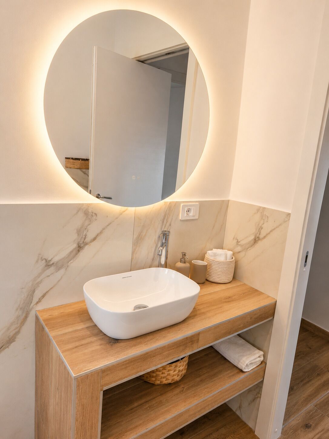 A bathroom with a white sink, wooden vanity, marble walls, and a round mirror with lighting.