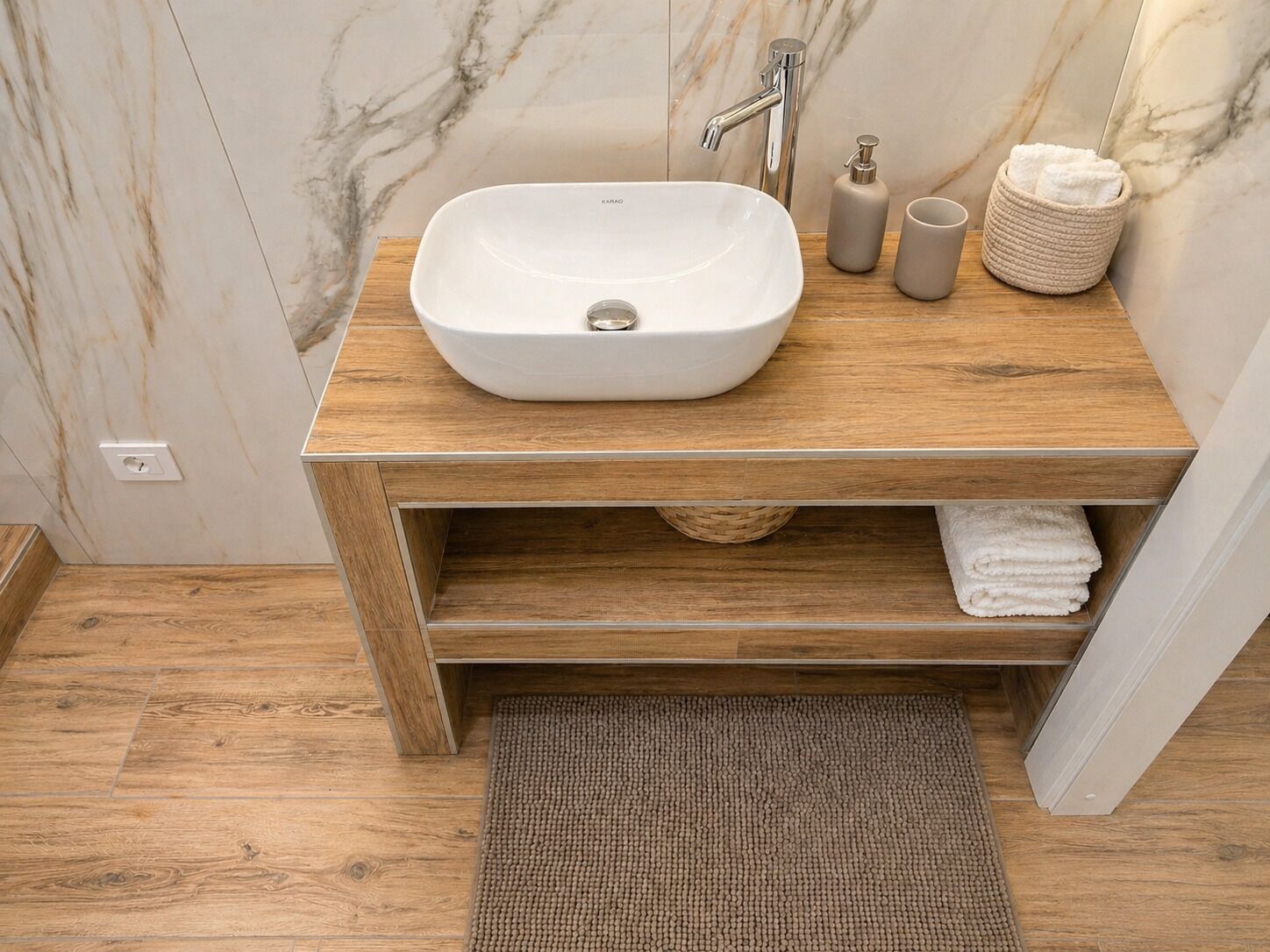 A bathroom with a white sink, wooden vanity, marble walls, and a woven mat.