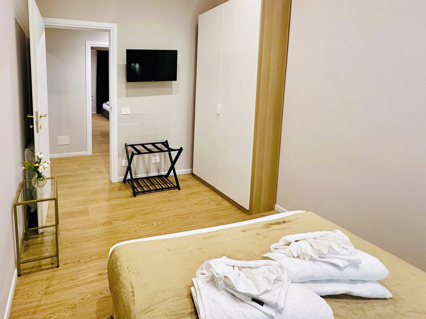 A hotel room with a bed, a TV mounted on the wall, a small black rack, and a wooden floor.