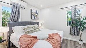 A modern bedroom with a large bed, a bedside table with a lamp, a potted plant, and a window with curtains.