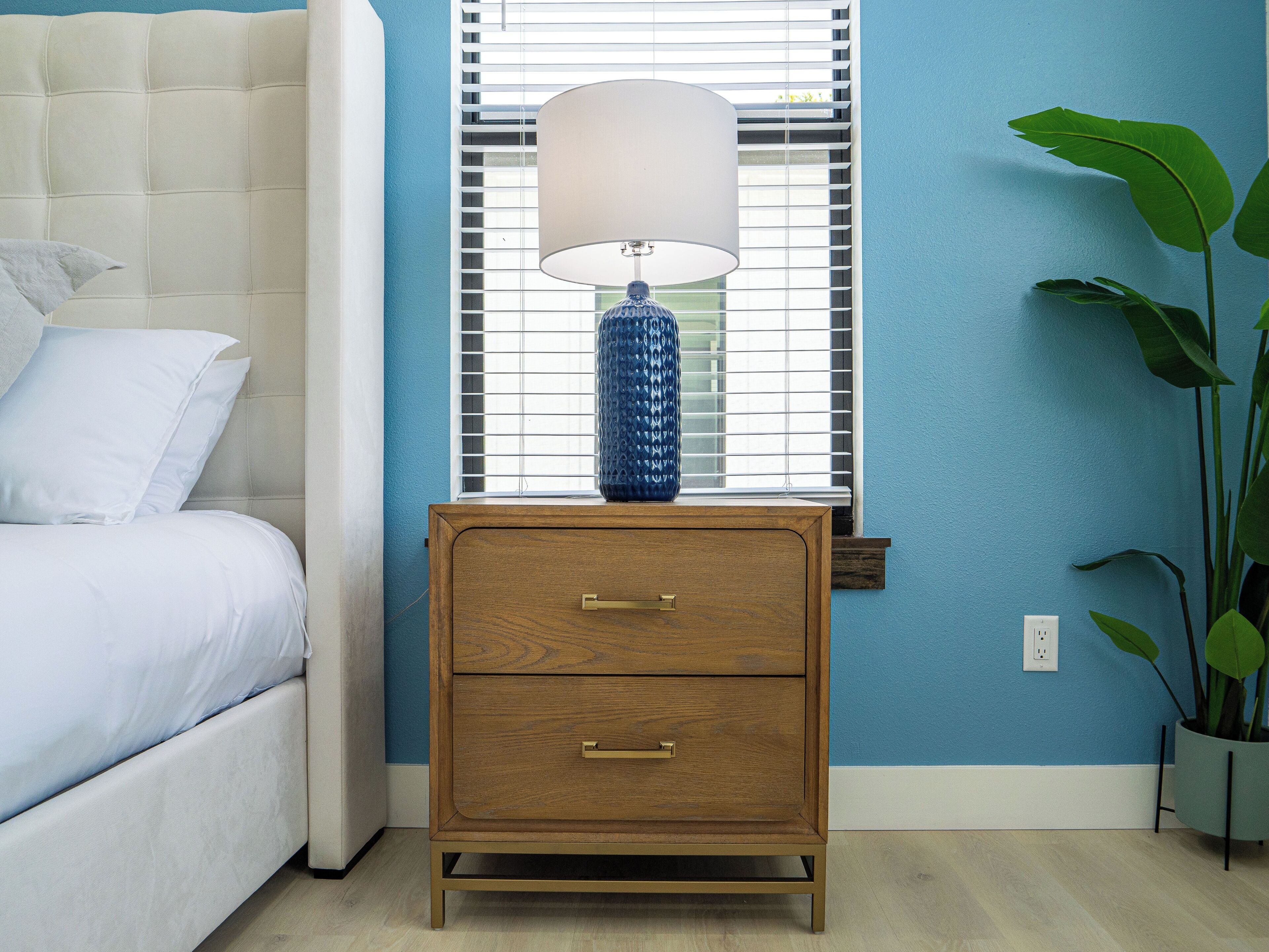 A bedroom with a bed, a nightstand with a lamp, a window with blinds, and a potted plant.