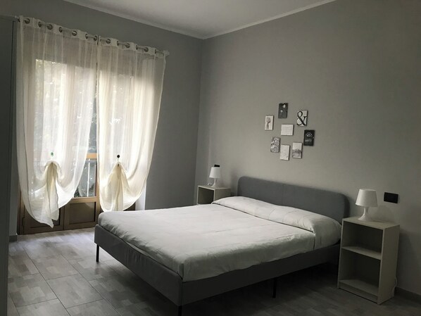 A bedroom with a bed, two bedside tables, a nightstand, a window with sheer curtains, and wall art.