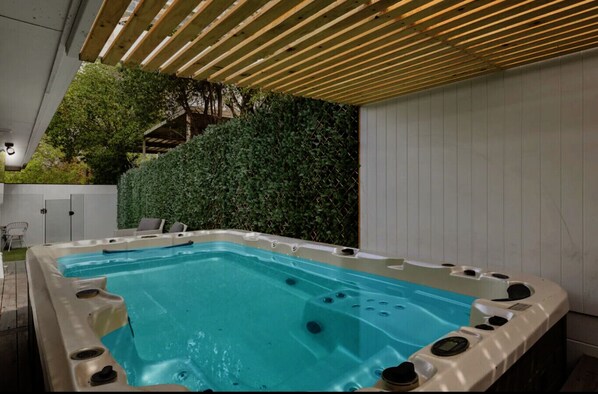 An outdoor hot tub area with a wooden pergola overhead, surrounded by greenery and a white wall.