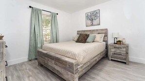 Un dormitorio con una cama de madera, una mesita de noche, una ventana con cortinas y un cuadro colgado en la pared.