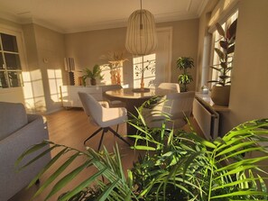 A modern living room with a dining table, chairs, and a sofa. There are several potted plants and a hanging light fixture. - Exklusives Ferienhaus am Waldrand – Veluwe entdecken und relaxen (Barneveld)