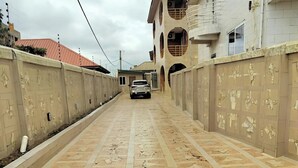 A paved walkway with a patterned design, flanked by walls with relief sculptures, leading to a building with multiple balconies and a car parked in the distance. - Charming 1-bedroom apartment in Tema with AC (Tema)
