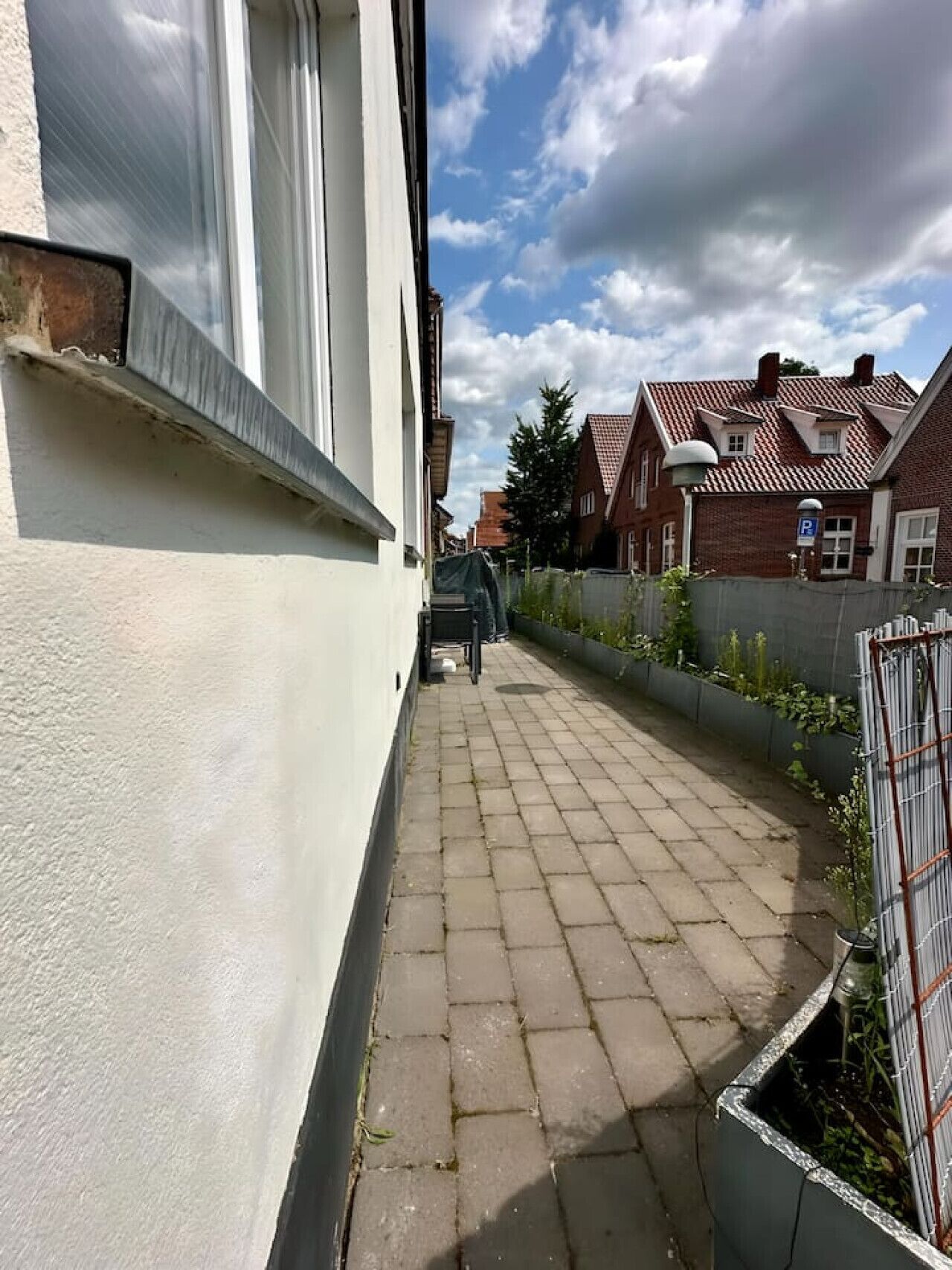 Ein schmaler gepflasterter Weg führt zu einem Wohngebäude mit weißer Fassade und Ziegelverkleidung.