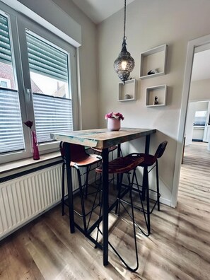 A dining area with a wooden table, black metal chairs, a hanging light fixture, and a window with blinds.