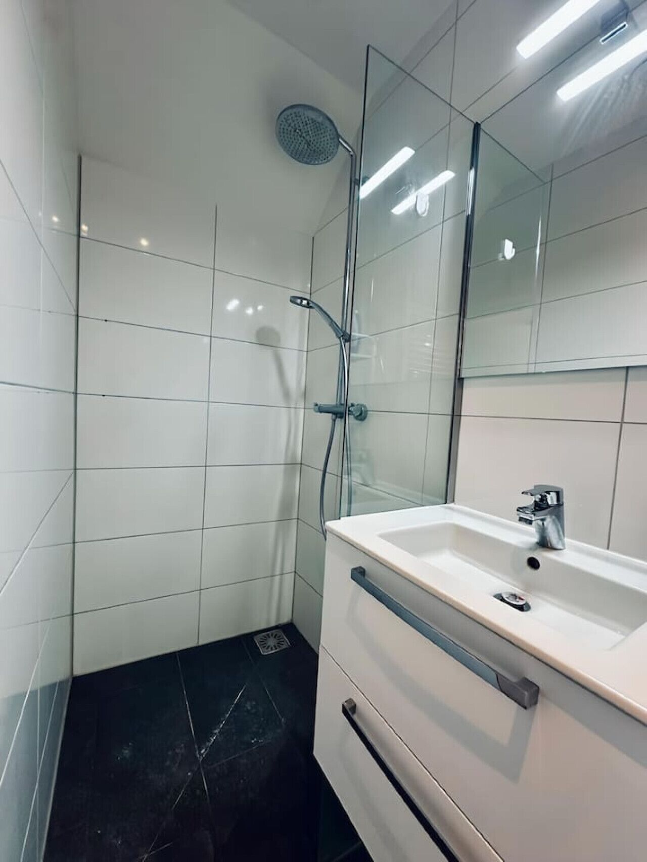 Een moderne badkamer met een glazen douchecabine, een wit wastafelmeubel met een spiegel erboven en een zwarte betegelde vloer.