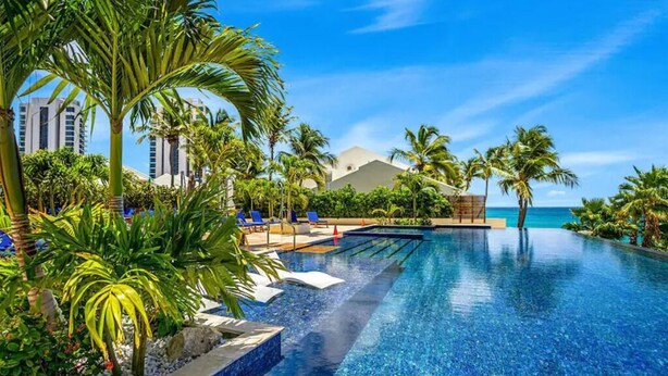 Una piscina infinita azul cristalina con palmeras y sillas de descanso, rodeada por un entorno de resort con vistas al océano.