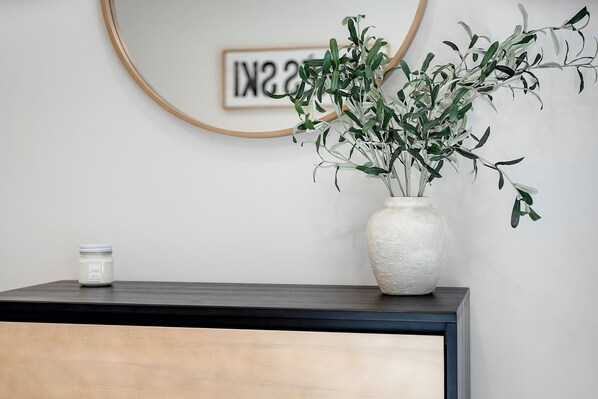 A minimalist room with a dark wooden console table, a white vase with green foliage, and a framed text on the wall.