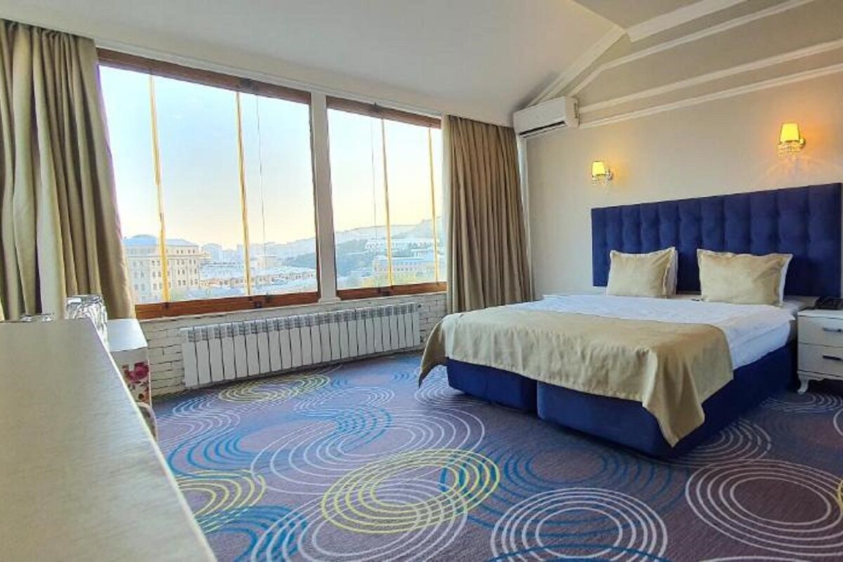 Hotellihuone, jossa on suuri sänky, yöpöytä, ikkuna verhoilla ja näkymä kaupunkiin.