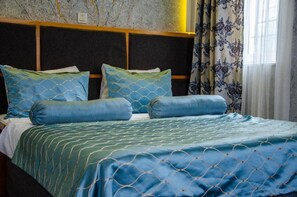 Un lit avec une couvre-lit à motifs bleus et verts, des coussins assortis, une tête de lit sombre et des rideaux à motifs floraux.