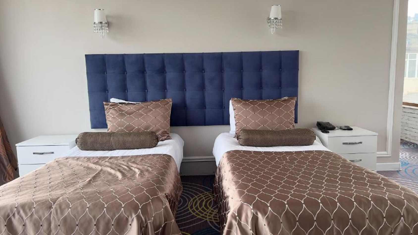 Een hotelkamer met twee bedden, een groot gestoffeerd hoofdeinde, nachtlampjes en een klein tafeltje met een telefoon.