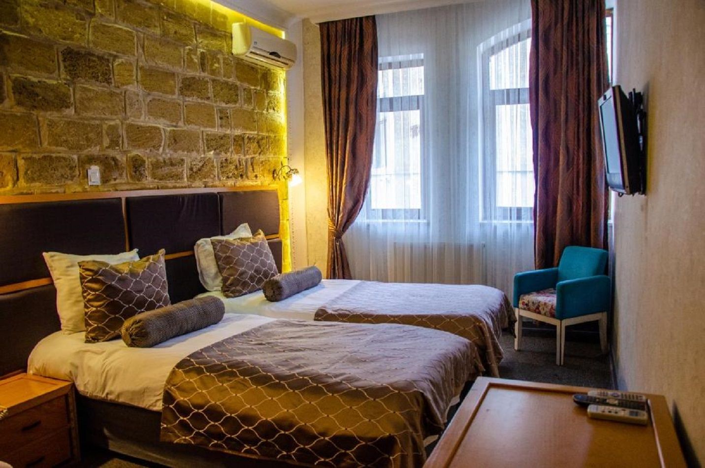 Une chambre d’hôtel avec deux lits, un mur en pierre, un climatiseur, une télévision, une chaise et une table avec des télécommandes.