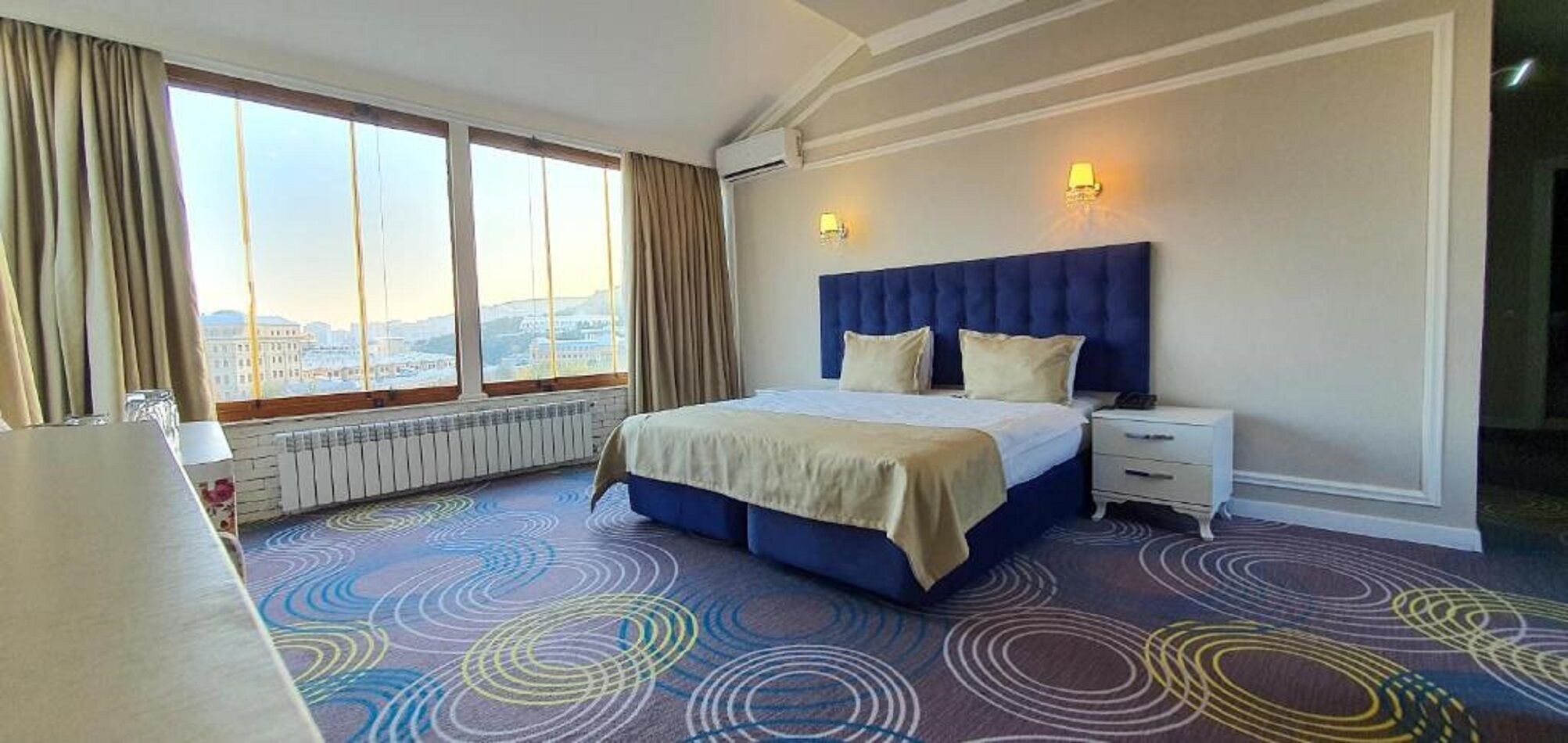 Een hotelkamer met een groot bed, een nachtkastje, een raam met gordijnen en uitzicht op de stad.