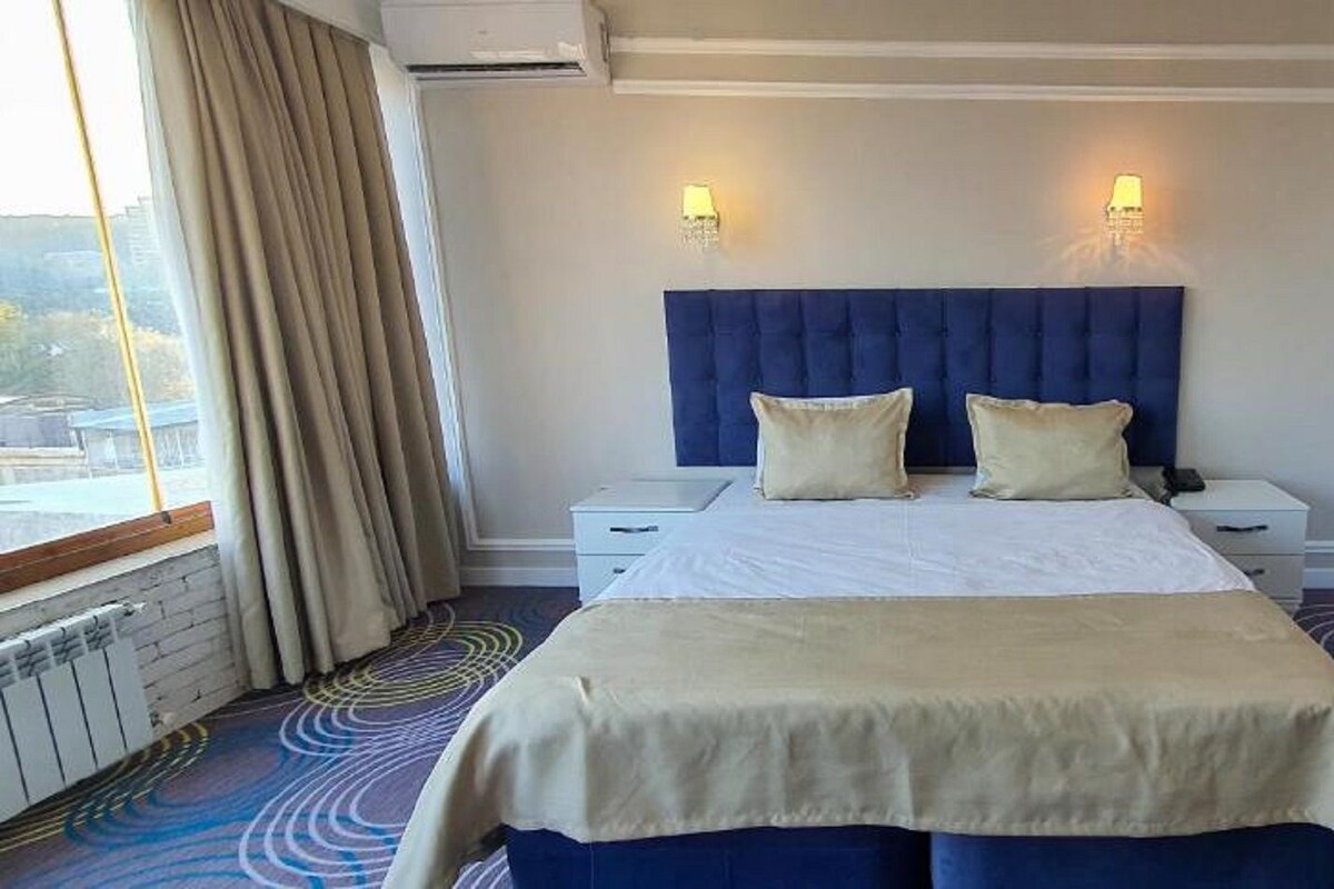 Une chambre d’hôtel avec un grand lit, deux tables de chevet, une tête de lit et une fenêtre avec des rideaux.