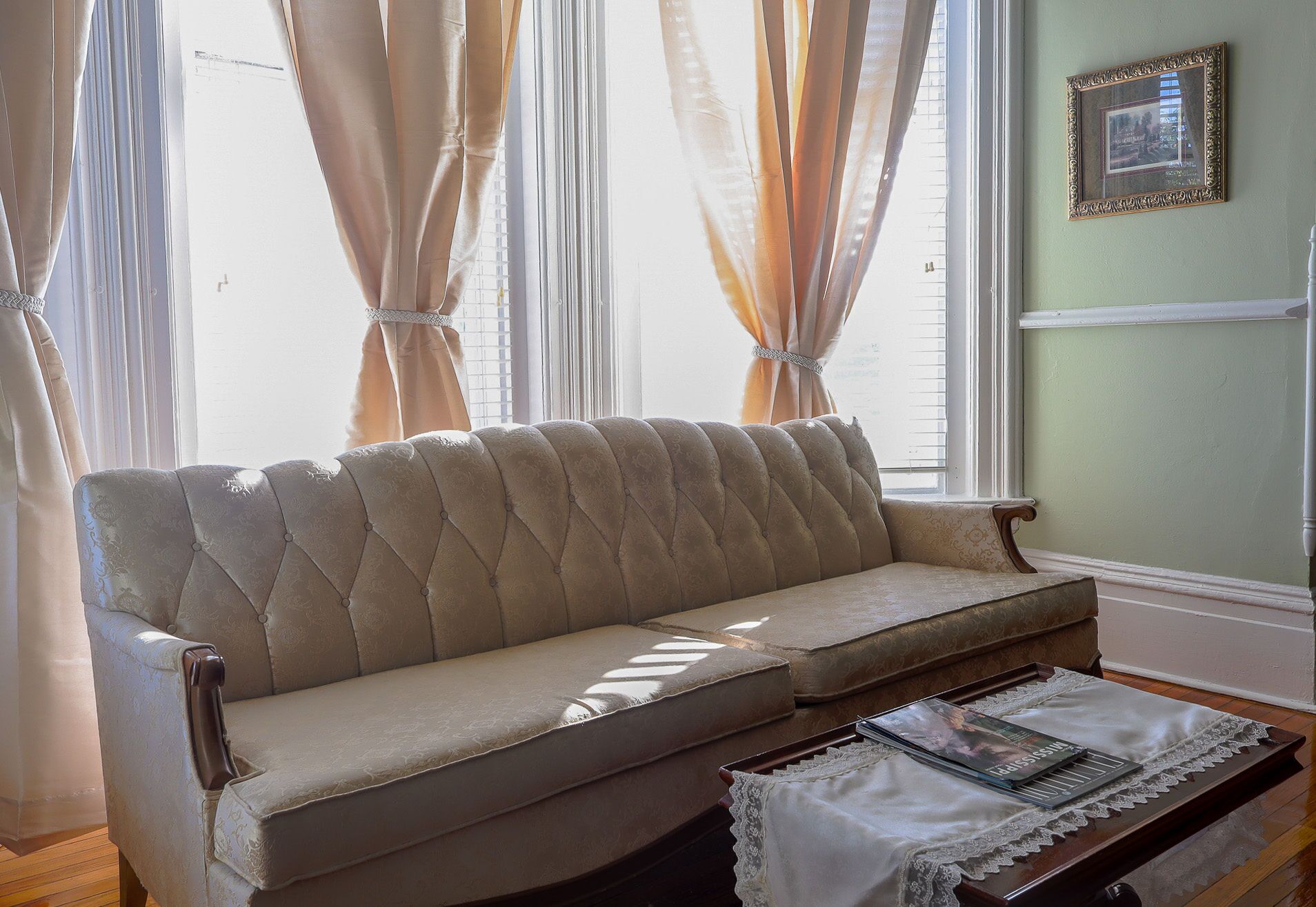 A beige sofa with a cushioned backrest and armrests, positioned in front of a window with sheer curtains.