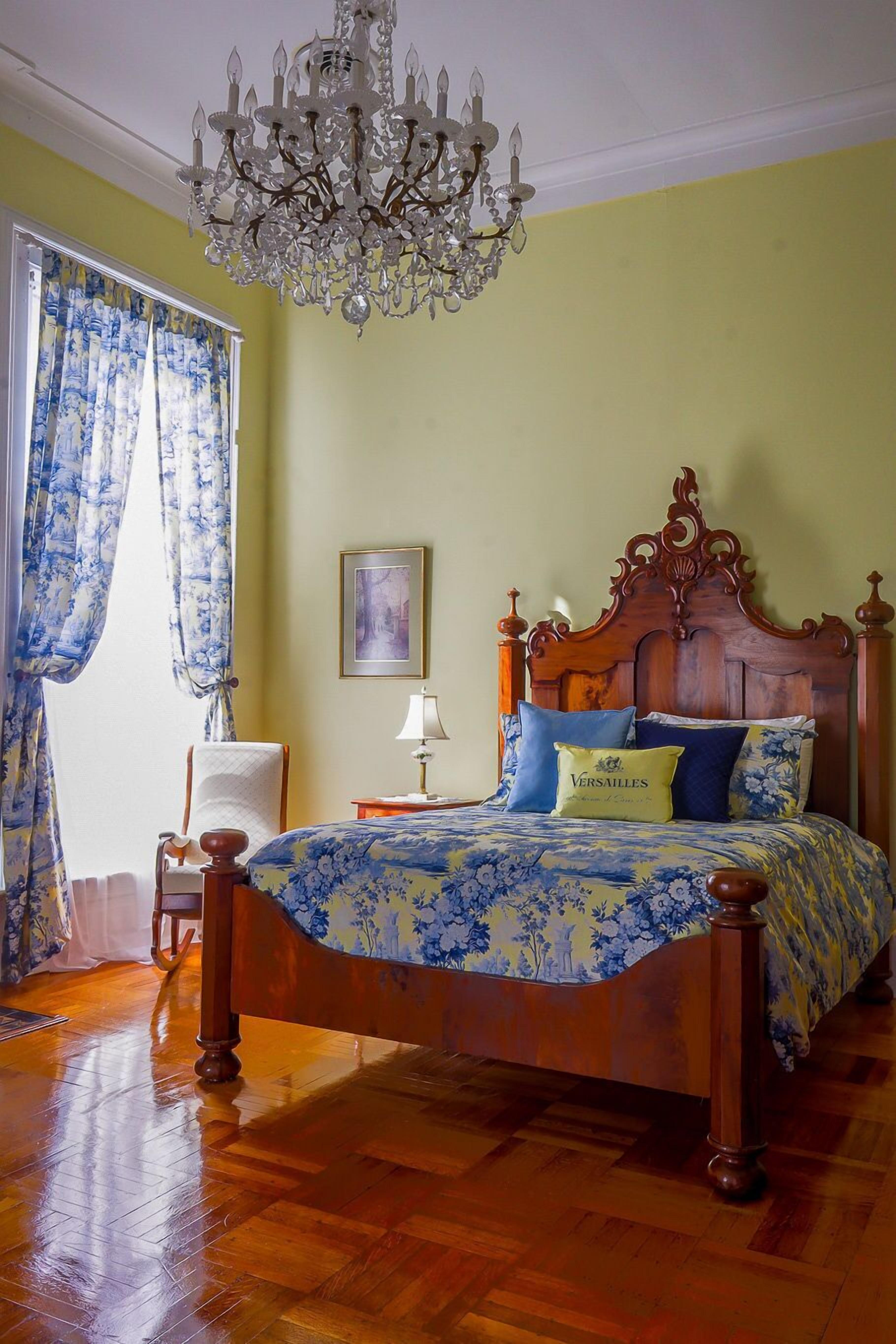A bedroom with a wooden bed, a chandelier, a chair, and a window with curtains.