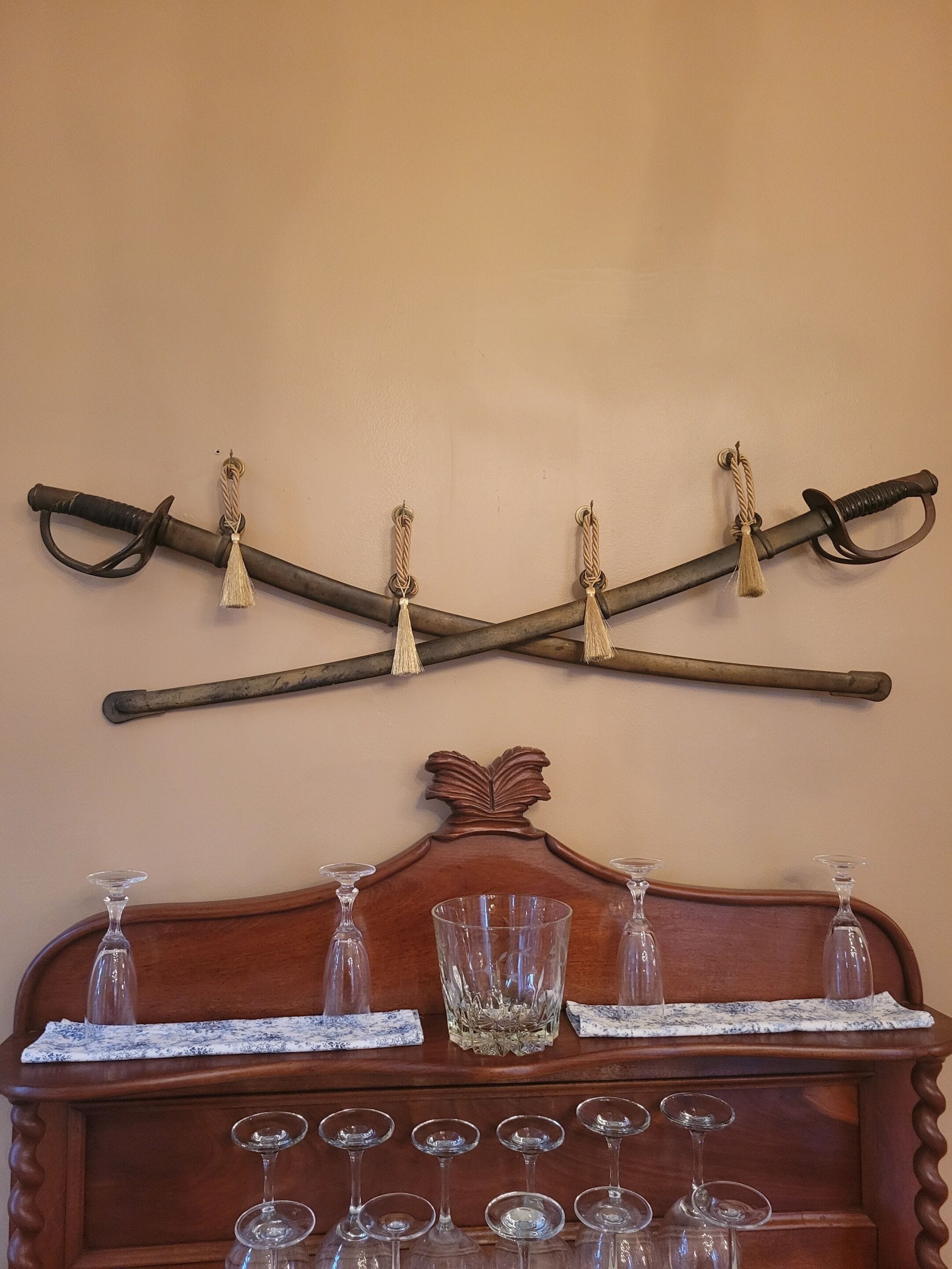 A wooden bar with a decorative headboard, a mounted sword with tassels, and a set of wine glasses arranged on the bar.