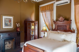 Un dormitorio con cama, cómoda de madera, espejo, chimenea y ventana con cortinas.