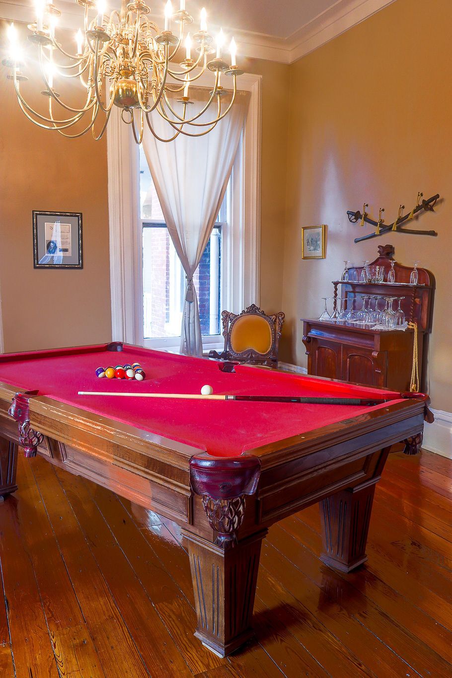A pool table with balls, a chandelier, a window with curtains, and a wooden cabinet with glasses.