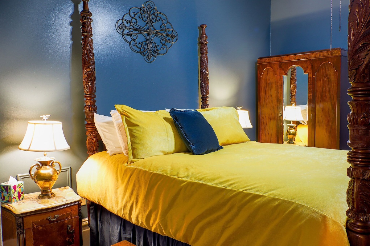 Een bed met een geel sprei, een houten hoofdbord, een nachtkastje met een lamp en een spiegel.