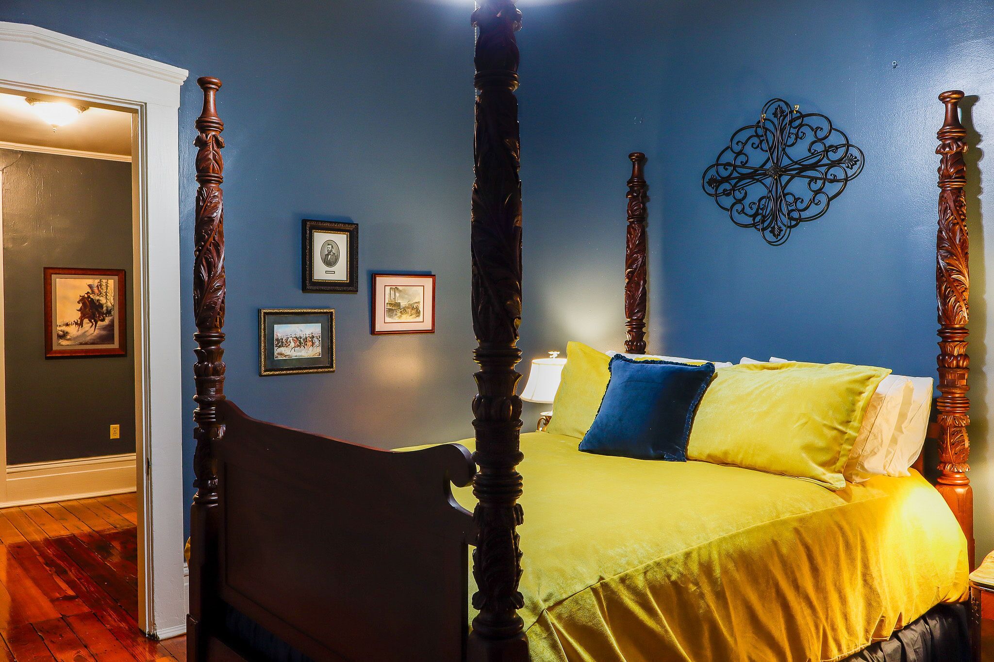 A four-poster bed with a yellow bedspread and blue pillows, a wooden floor, and framed pictures on the wall.