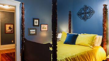 A four-poster bed with a yellow bedspread and blue pillows, a wooden floor, and framed pictures on the wall.