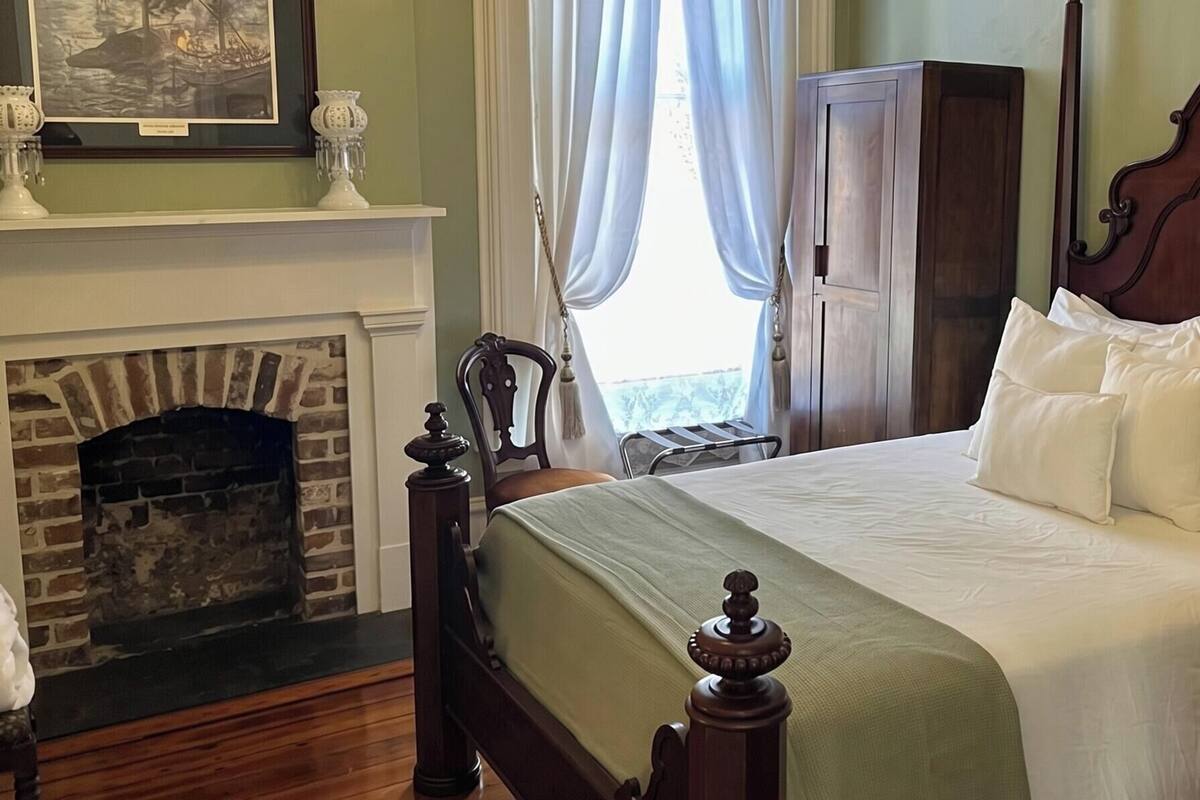 A bedroom with a bed, a wooden headboard, a nightstand, a dresser, a fireplace, and a window with curtains.