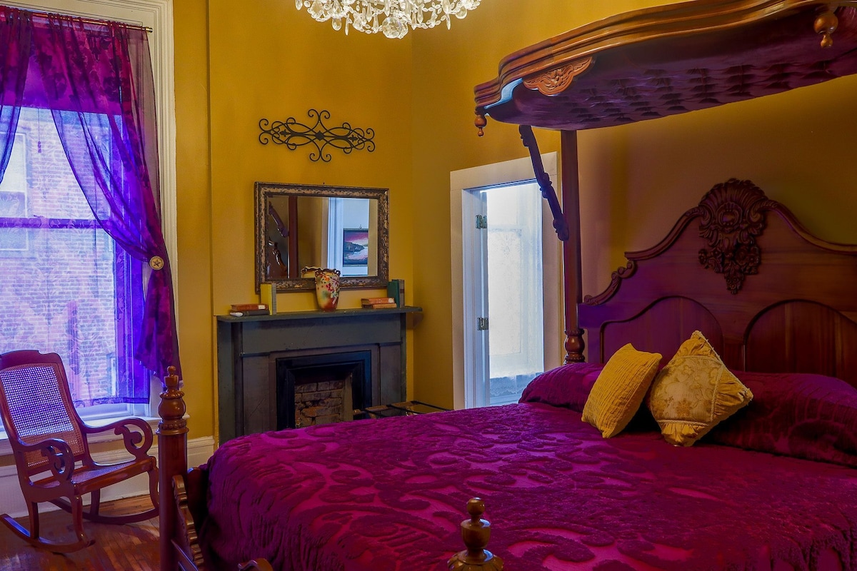 Een kamer met een hemelbed, een houten schommelstoel, een open haard en een grote spiegel aan de muur.