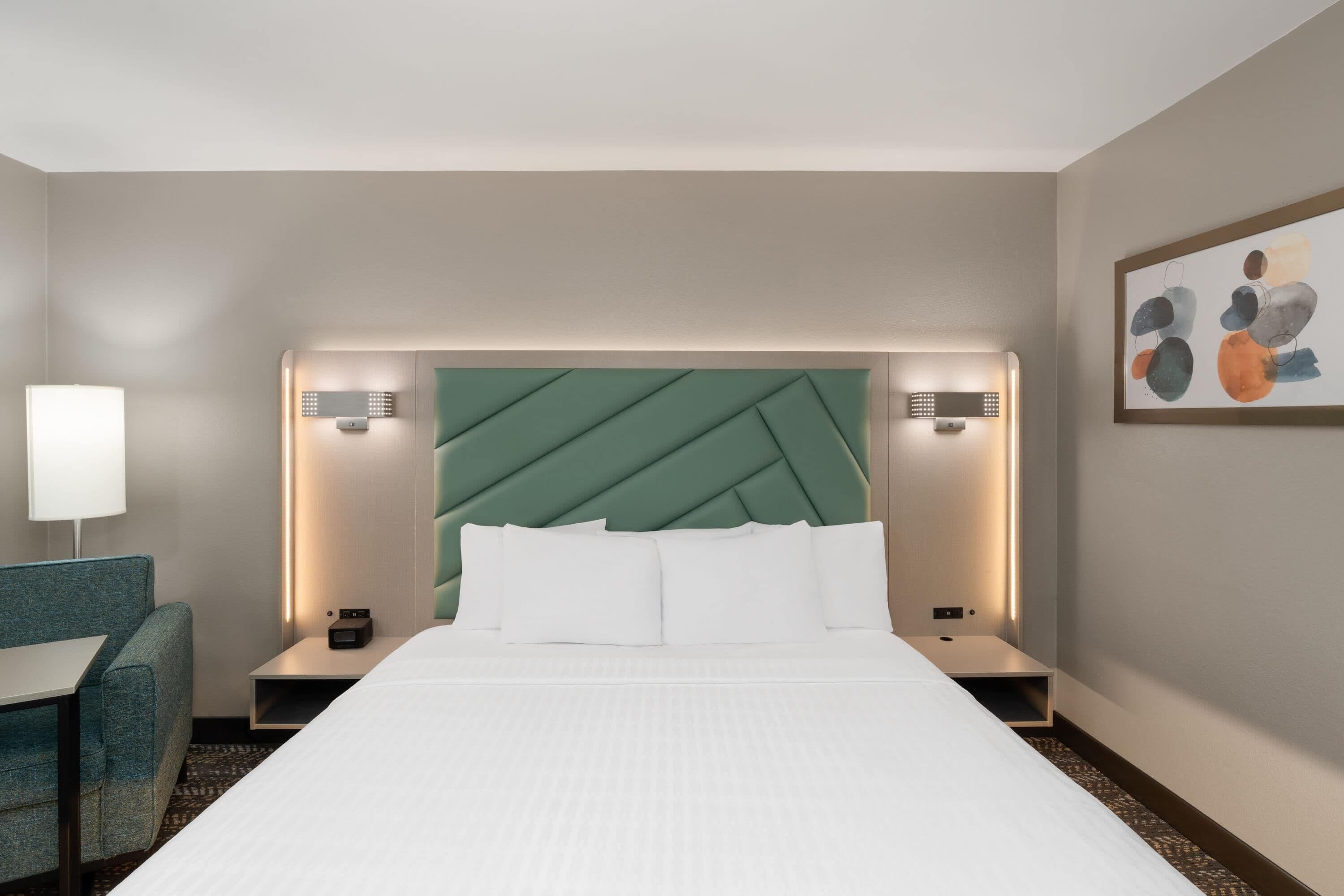 A hotel room with a bed, a desk, a chair, a nightstand, and a wall-mounted artwork.
