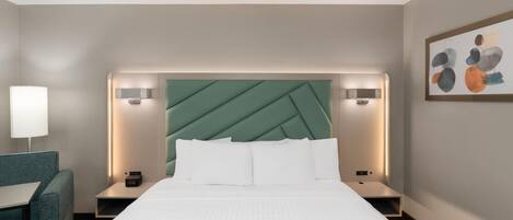 A hotel room with a bed, a desk, a chair, a nightstand, and a wall-mounted artwork.