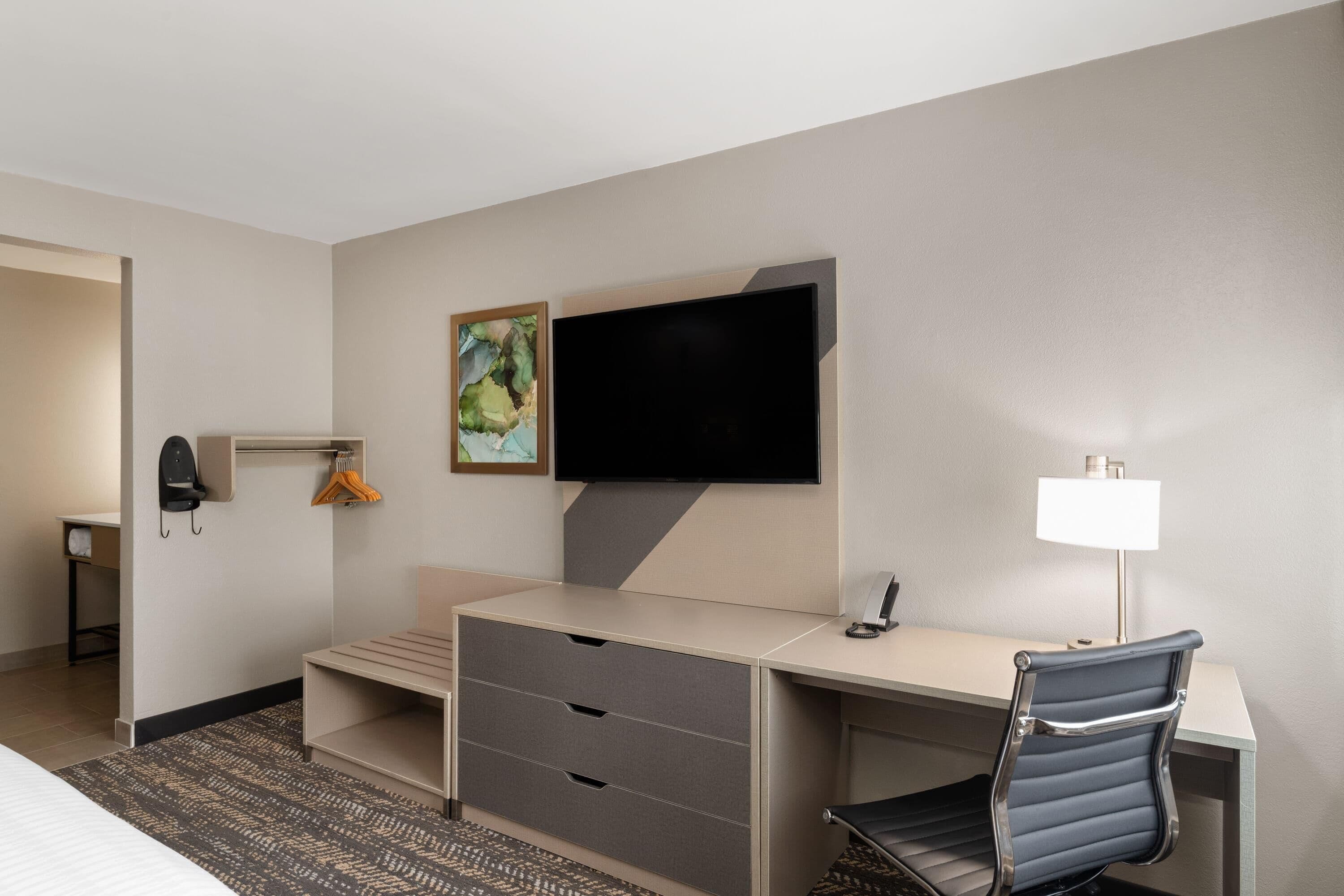 A hotel room with a bed, a desk with drawers, a television mounted on the wall, a chair, a lamp, and a coat rack with hangers.