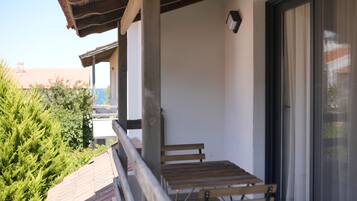 Ein Balkon mit Holztisch und -bänken, einer Schiebetür und Blick auf Grünflächen sowie Gebäude im Hintergrund.