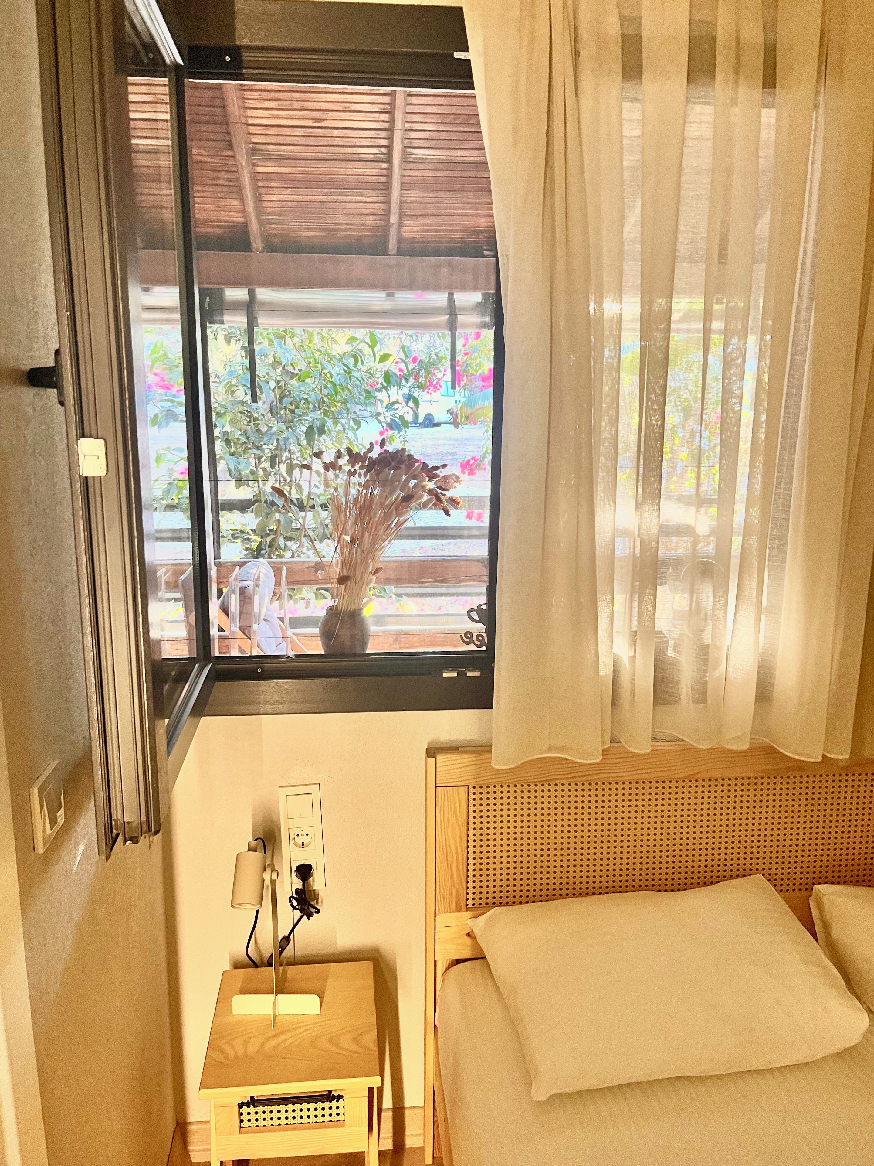 Ein Zimmer mit einem Bett, einem kleinen Tisch mit einer Lampe, einer Steckdose und einem Fenster mit Blick auf Grünflächen und ein parkendes Auto.