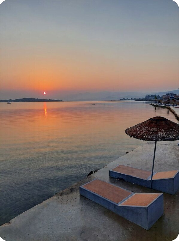 Zonsondergang boven kalm water, met een betonnen looppad en een rieten parasol.