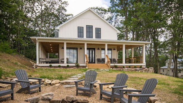 Een groot wit huis met een overdekte veranda, veel ramen en een vuurkorf met Adirondack-stoelen ervoor.