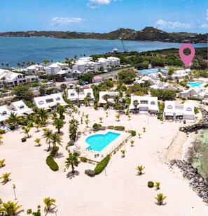 Une maison de vacances située sur la plage, avec une piscine, des bâtiments blancs et une vue dégagée sur l’océan.