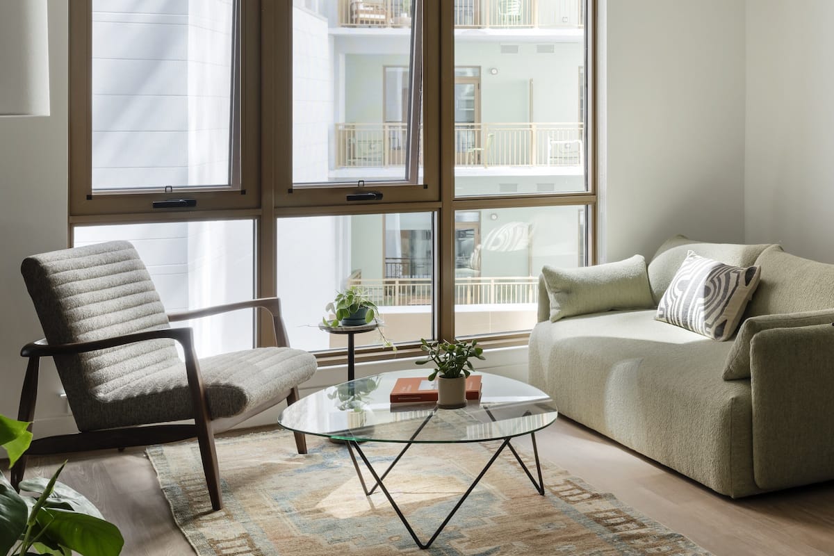 A modern living room with a glass coffee table, a grey armchair, a beige sofa, and large windows offering a view of a residential building.