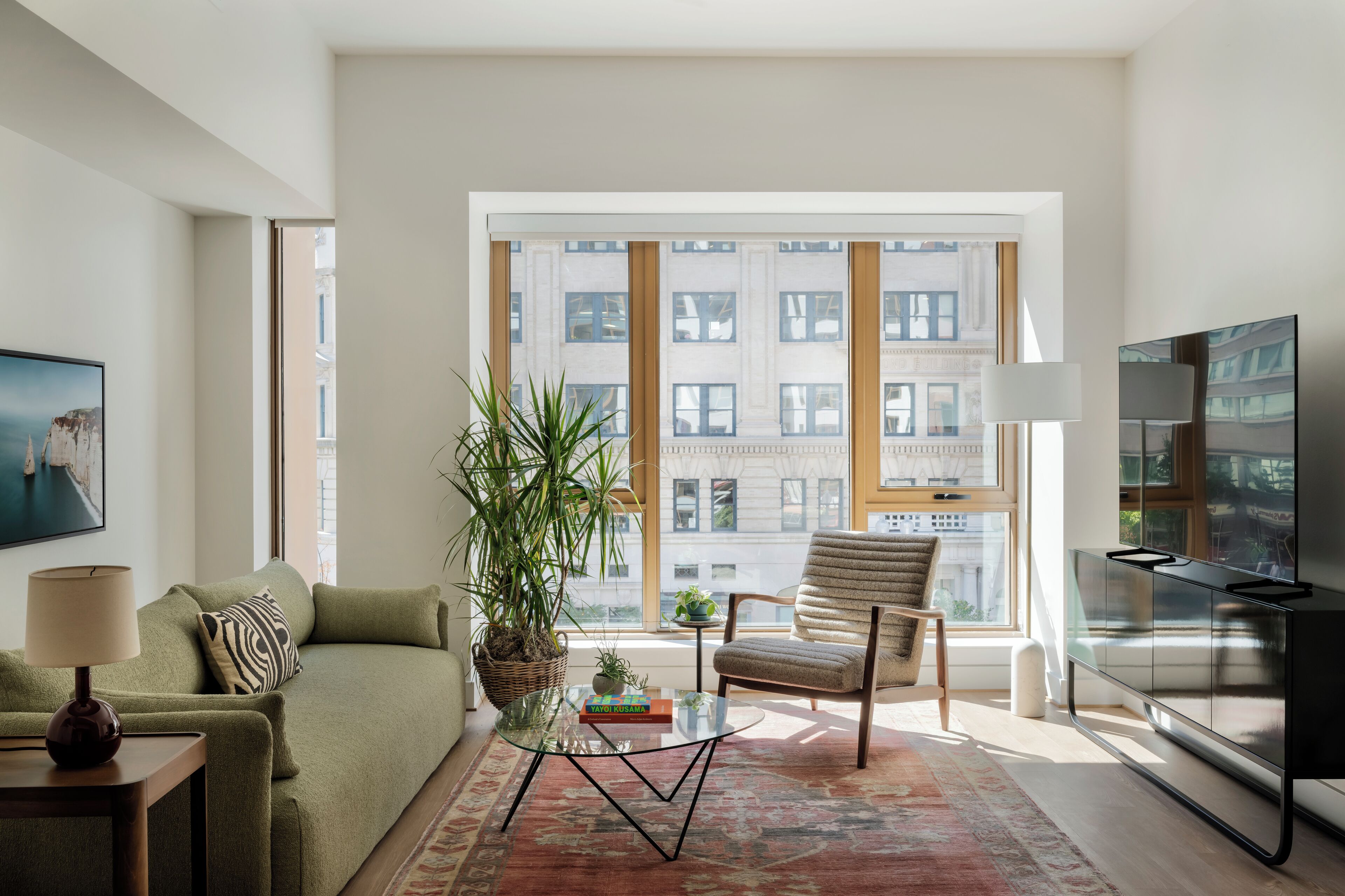 A modern living room with a sofa, a glass coffee table, a chair, a TV, and a large window with a city view.