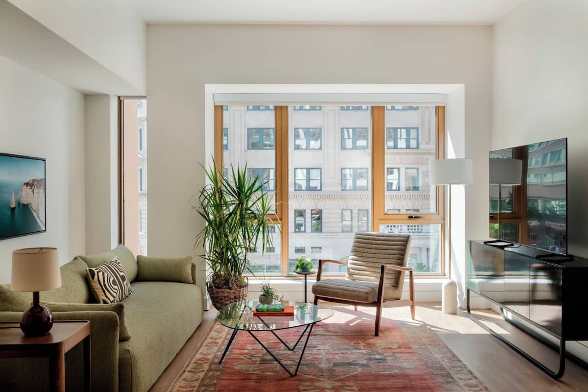 A modern living room with a sofa, armchair, TV, and large windows offering a city view.