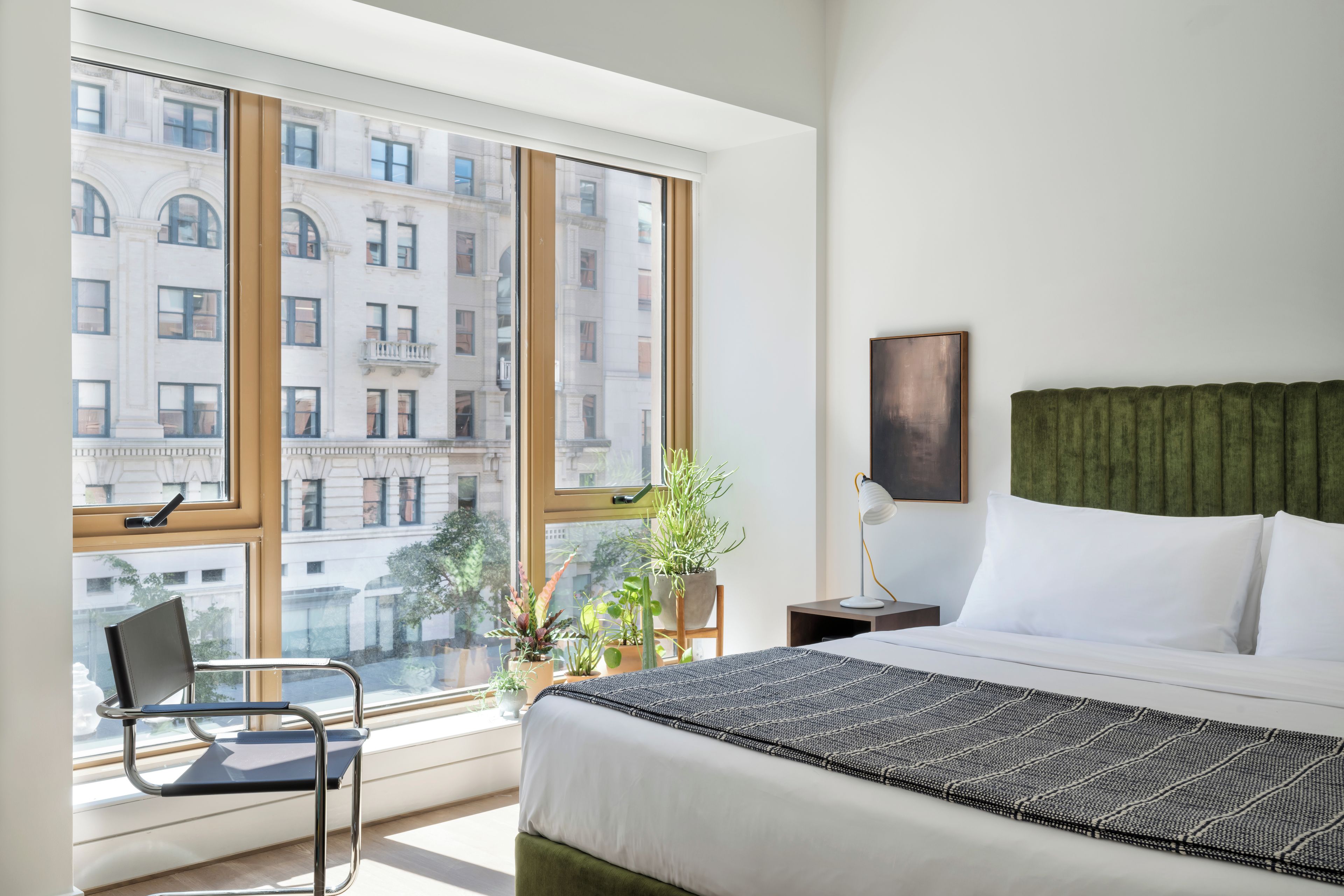 A hotel room with a large window, a bed with white linens, a green upholstered headboard, a bedside table with a lamp, and a chair.
