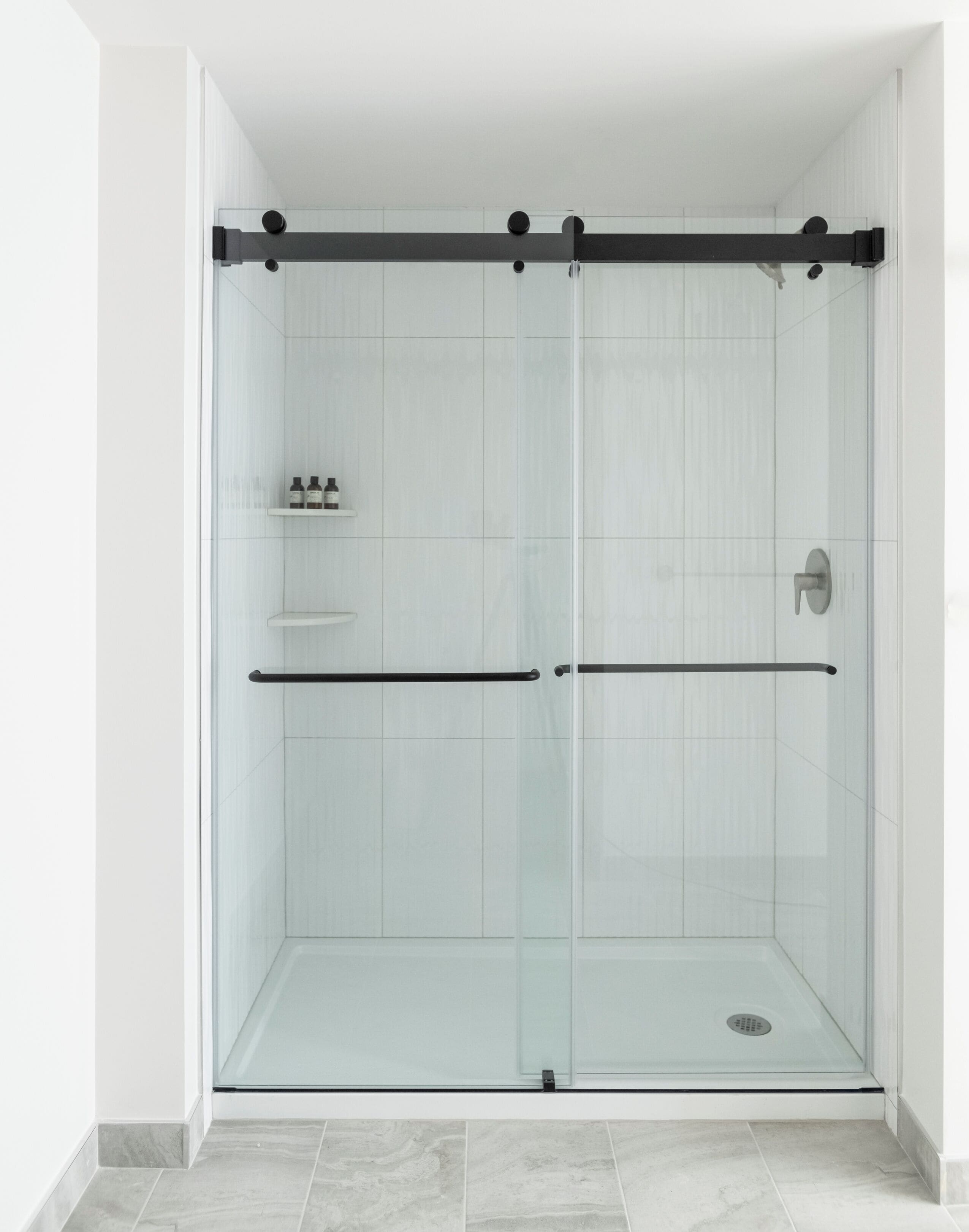 A glass-enclosed shower with a black handle and a shelf with toiletries.