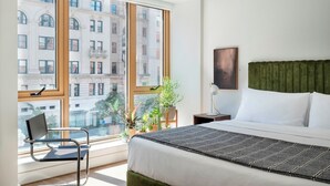 A hotel room with a large window, a bed with white linens, a green upholstered headboard, a bedside table with a lamp, and a chair by the window.
