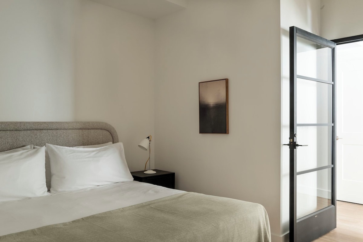 A bedroom with a bed, a bedside table with a lamp, a framed picture on the wall, and a sliding glass door leading to a balcony.
