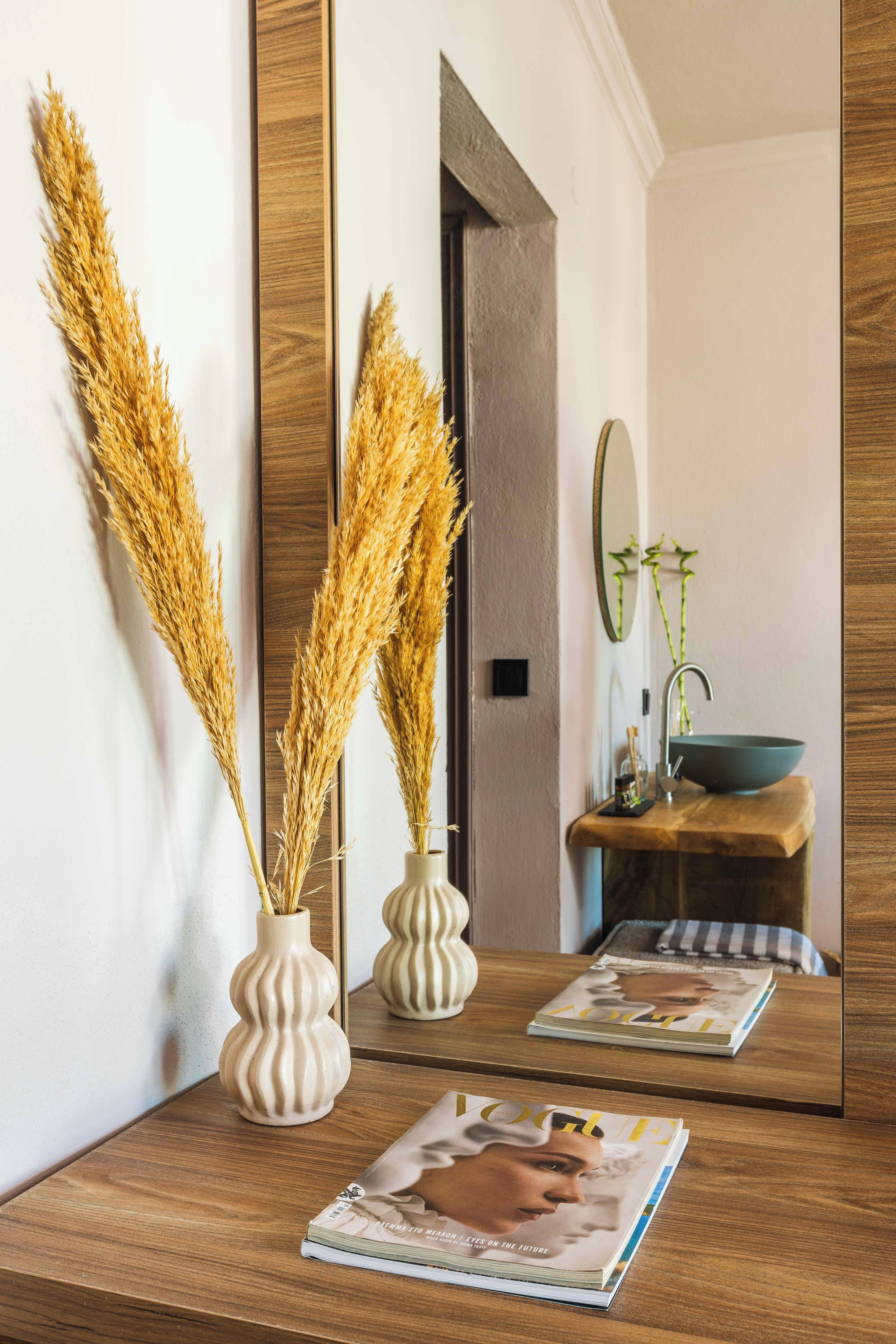 A wooden vanity with a mirror, two vases with dried plants, a sink, and a magazine on the counter.
