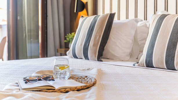 A neatly made bed with striped pillows, a glass of water with a lemon slice, and a pair of glasses on a woven tray. - Casa Blue Hotel (Skiathos)