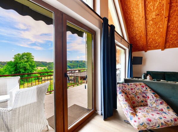 A balcony with a view of green hills, a wicker chair, a sofa with floral upholstery, and wooden paneling on the ceiling.