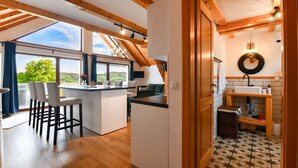 A modern kitchen with a central island, wooden beams, and large windows offering a view of greenery.